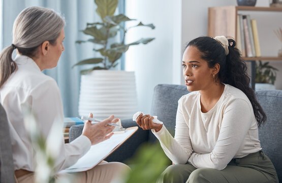 Two women sitting in a comfortable living room setting during a counseling session, with one taking notes on a clipboard while the other speaks and gestures thoughtfully.